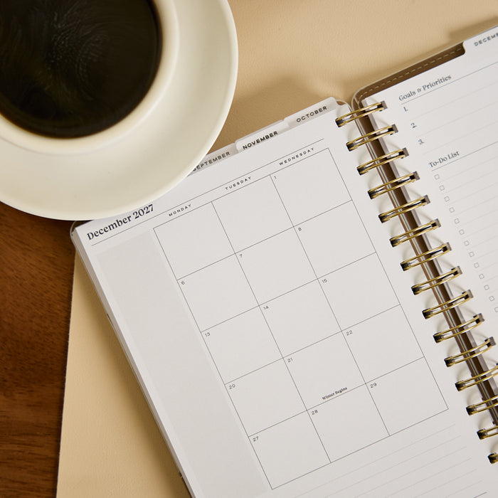 2027 Ivory Delicate Dots Customizable Spiral Citrine Planner - Small Professional Layout - 12 Months open to a December 2027 monthly calendar spread with spiral binding beside coffee