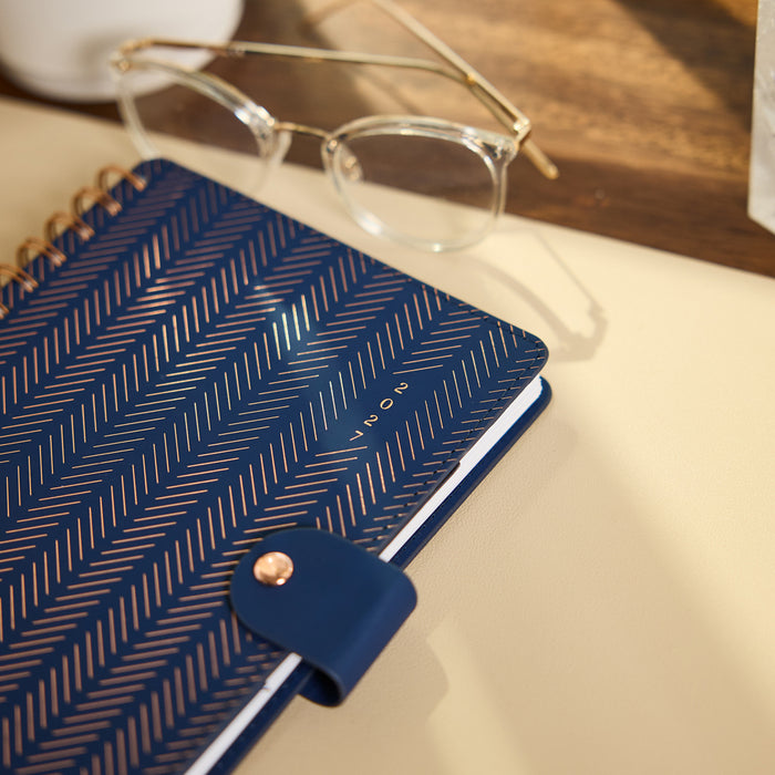 2027 Navy Herringbone Customizable Spiral Citrine Planner - Small Professional Layout - 12 Months with navy cover and rose gold spiral binding beside eyeglasses on a desk