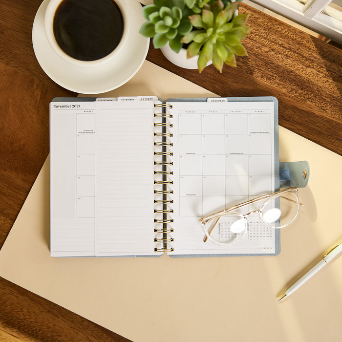2027 Pastel Horizon Customizable Spiral Citrine Planner - Small Professional Layout - 12 Months open to a monthly spread with spiral binding and glasses nearby