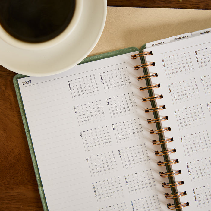 2027 Sage Waves Customizable Spiral Citrine Planner - Small Professional Layout - 12 Months open to monthly calendar pages beside a cup of coffee on a wooden desk