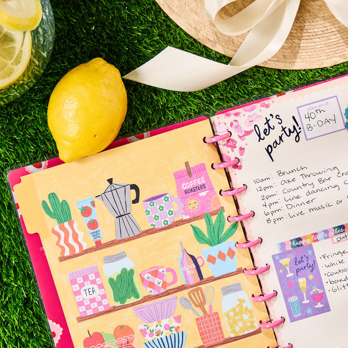 Open Happy Little Vibes - Dotted Lined Classic Notebook - 60 Sheets showing colorful kitchen-themed illustrations on left page and a handwritten party schedule on right page with pink discs binding the notebook.
