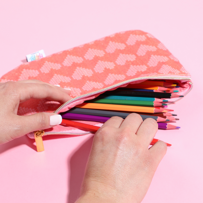 Person’s hands opening a coral patterned Miss Maker - Classic Banded Pen Pouch filled with colorful sharpened pencils on a pink background