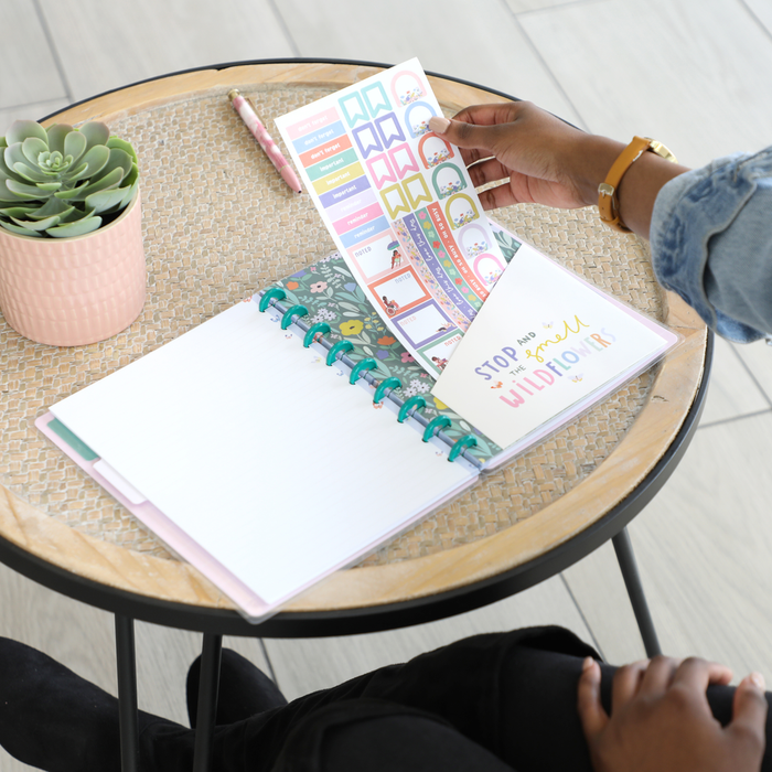 Person organizing colorful planner stickers into a Happy Planner notebook on a round table with a succulent, featuring the Squad Goals Love Every Season - Classic Accessory Pack.
