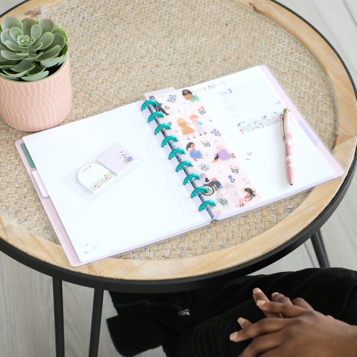 Open disc bound planner featuring translucent decorative cover with diverse illustrated characters, lined and dotted filler paper, floral stickers, and pink pen on a wicker tabletop alongside a potted succulent, showcasing Squad Goals Love Every Season - Classic Accessory Pack.