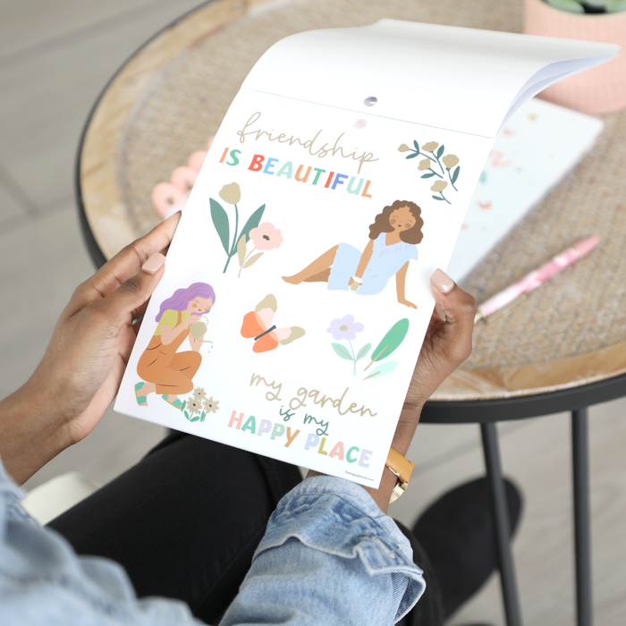 Person holding a sheet of colorful stickers featuring illustrated women, flowers, a butterfly, and phrases like "friendship is beautiful" and "my garden is my happy place" from the Squad Goals Love Every Season - Large Value Pack Stickers collection.