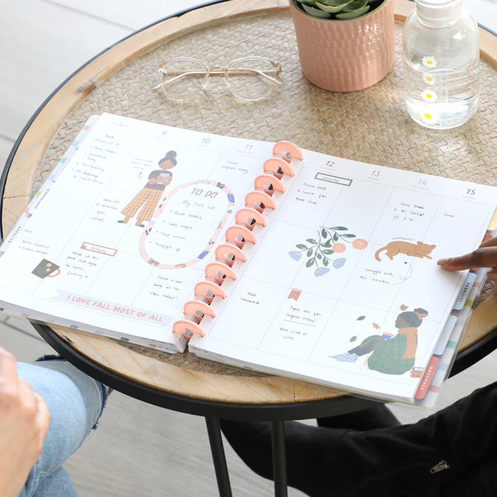 Open disc bound planner on round wicker table with vinyl rings, featuring illustrations of women and a dog, handwritten notes, and decorative stickers from the Squad Goals Love Every Season - Large Value Pack Stickers collection.