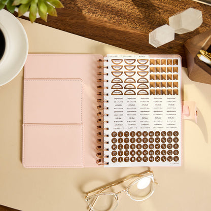 2027 Blush Diagonals Customizable Spiral Citrine Planner - Small Professional Layout - 12 Months open beside a coordinating sticker sheet on a desk