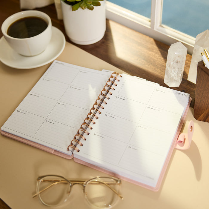2027 Blush Diagonals Customizable Spiral Citrine Planner - Small Professional Layout - 12 Months open on a desk showing a weekly layout beside coffee and glasses