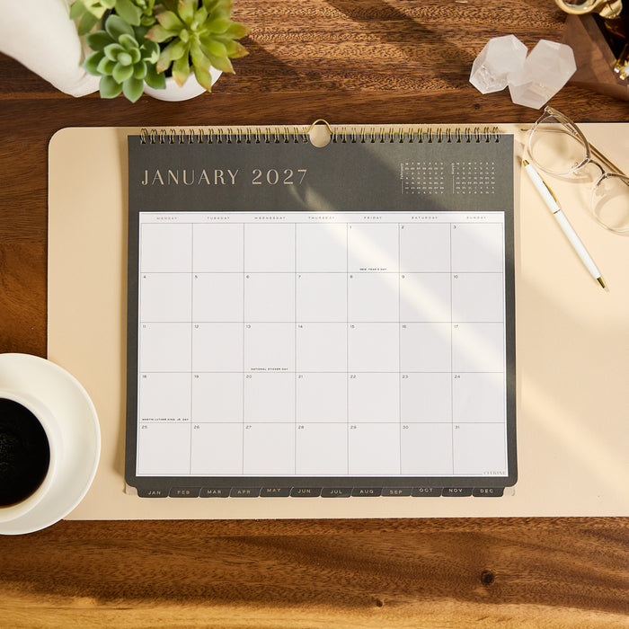 2027 Midnight Citrine Wall Calendar - Small - 12 Months displayed open to January 2027 on a desk beside a plant coffee glasses crystals and pen