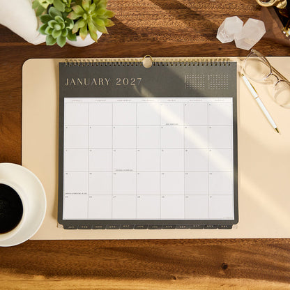 2027 Midnight Citrine Wall Calendar - Small - 12 Months displayed open to January 2027 on a desk beside a plant coffee glasses crystals and pen