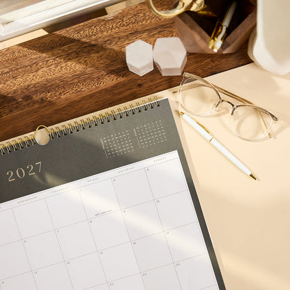 2027 Midnight Citrine Wall Calendar - Small - 12 Months open on a wooden desk with a monthly grid gold spiral binding glasses and a pen