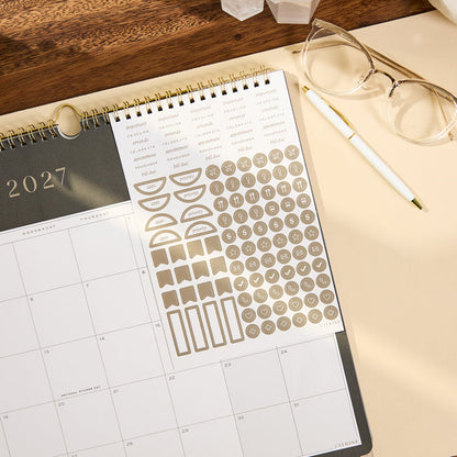 2027 Midnight Citrine Wall Calendar - Small - 12 Months open to a monthly layout with decorative stickers beside glasses and a pen on a wood desk
