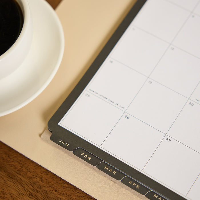 2027 Midnight Citrine Wall Calendar - Small - 12 Months on a desk with monthly tabs and a coffee cup nearby
