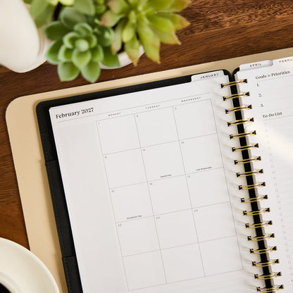 2027 Midnight Customizable Spiral Citrine Planner - Classic Professional Layout - 12 Months open to a February 2027 calendar with gold spiral binding on a wood desk