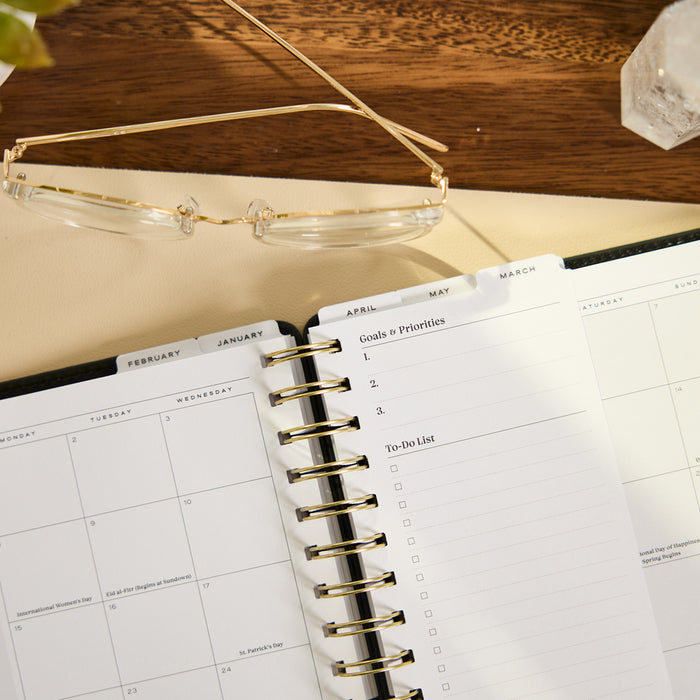 2027 Midnight Micro Dots Customizable Spiral Citrine Planner - Small Professional Layout - 12 Months open to a monthly calendar and goals and priorities checklist beside eyeglasses