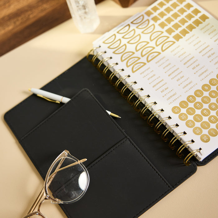 2027 Midnight Plaid Customizable Spiral Citrine Planner - Small Professional Layout - 12 Months open with gold spiral binding and planning stickers beside a black folio and glasses
