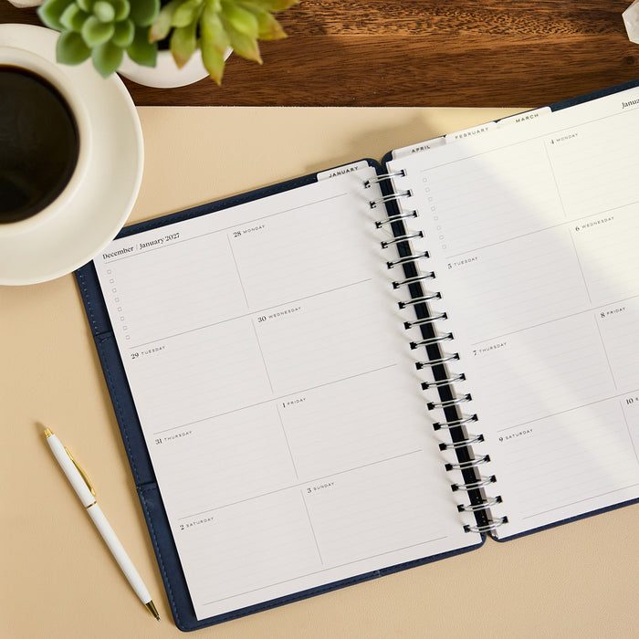 2027 Navy Customizable Spiral Citrine Planner - Classic Professional Layout - 12 Months open to a monthly spread with pen, coffee, and succulent nearby