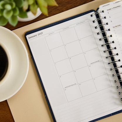 2027 Navy Customizable Spiral Citrine Planner - Small Professional Layout - 12 Months open to a January 2027 monthly calendar spread beside coffee and a succulent