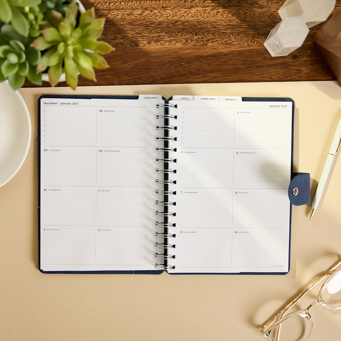 2027 Navy Customizable Spiral Citrine Planner - Small Professional Layout - 12 Months open to a monthly spread with dated writing spaces on a desk