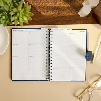 2027 Navy Customizable Spiral Citrine Planner - Small Professional Layout - 12 Months open to a monthly spread with dated writing spaces on a desk