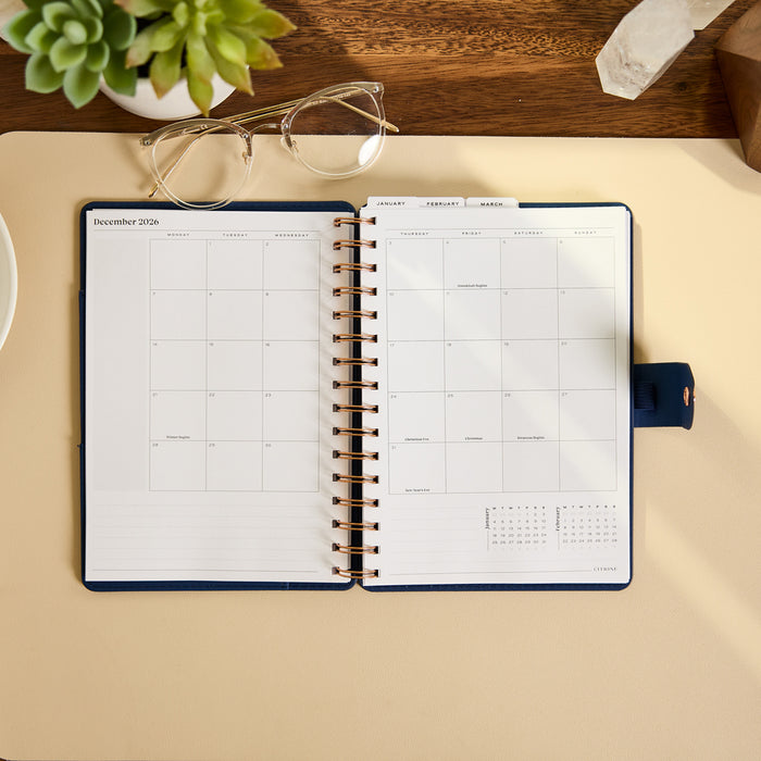 2027 Navy Herringbone Customizable Spiral Citrine Planner - Small Professional Layout - 12 Months open to a monthly calendar spread on a desk