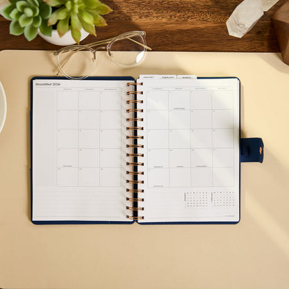 2027 Navy Herringbone Customizable Spiral Citrine Planner - Small Professional Layout - 12 Months open to a monthly calendar spread on a desk