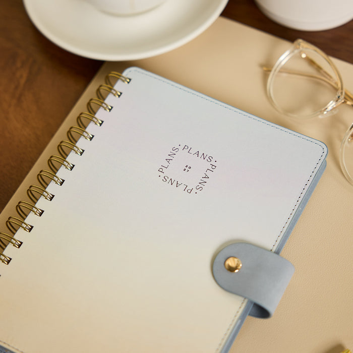 2027 Pastel Horizon Customizable Spiral Citrine Planner - Small Professional Layout - 12 Months with gold spiral binding and gray strap closure on a desk beside glasses and a cup