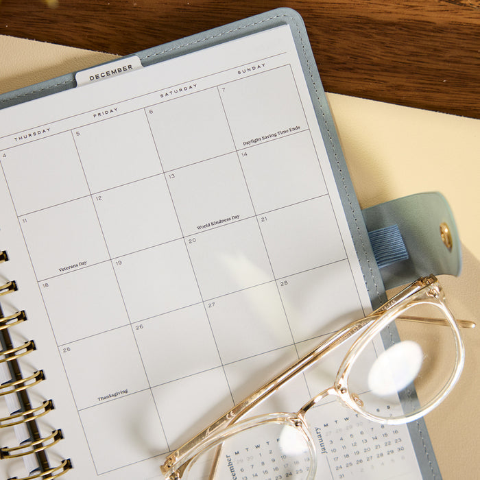 2027 Pastel Horizon Customizable Spiral Citrine Planner - Small Professional Layout - 12 Months open to a December calendar with glasses on a wooden desk