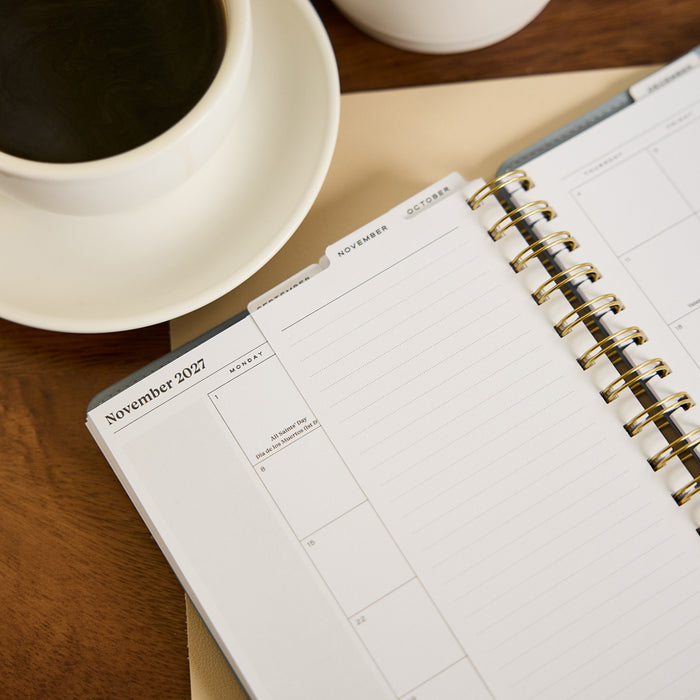 2027 Pastel Horizon Customizable Spiral Citrine Planner - Small Professional Layout - 12 Months open to a November 2027 monthly spread beside a cup of coffee