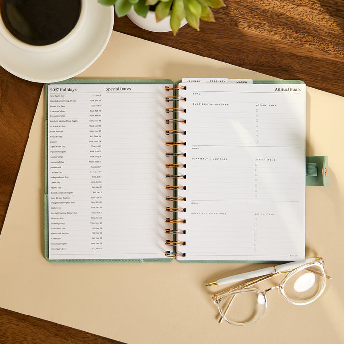 2027 Sage Waves Customizable Spiral Citrine Planner - Small Professional Layout - 12 Months open to holiday planning and annual goals on a desk beside coffee and glasses