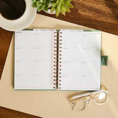 2027 Sage Waves Customizable Spiral Citrine Planner - Small Professional Layout - 12 Months open to a monthly spread on a desk beside coffee and glasses