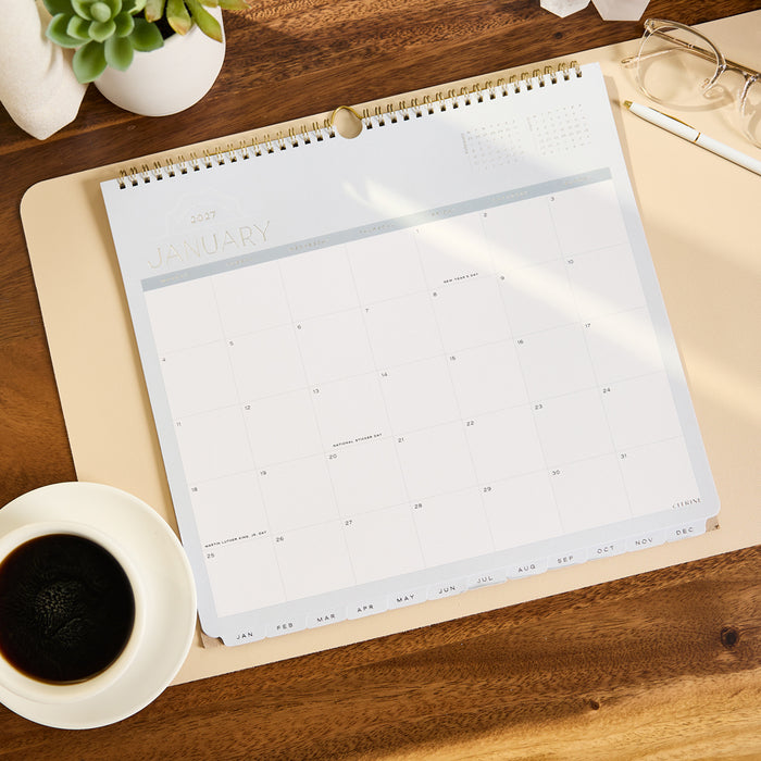 2027 Soft Frame Citrine Wall Calendar - Small - 12 Months open to a January monthly calendar on a wood desk beside coffee and glasses