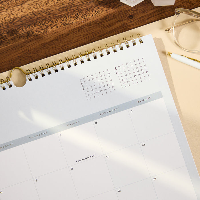 2027 Soft Frame Citrine Wall Calendar - Small - 12 Months open to a monthly grid with gold spiral binding on a wooden desk