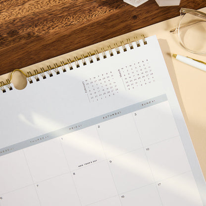 2027 Soft Frame Citrine Wall Calendar - Small - 12 Months open to a monthly grid with gold spiral binding on a wooden desk