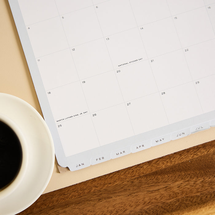 2027 Soft Frame Citrine Wall Calendar - Small - 12 Months displayed open on a wooden desk beside coffee with monthly dates and tabbed months