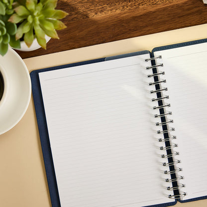 Citrine - Navy - Classic Customizable Spiral Notebook open to blank lined pages on a desk beside a plant and coffee cup