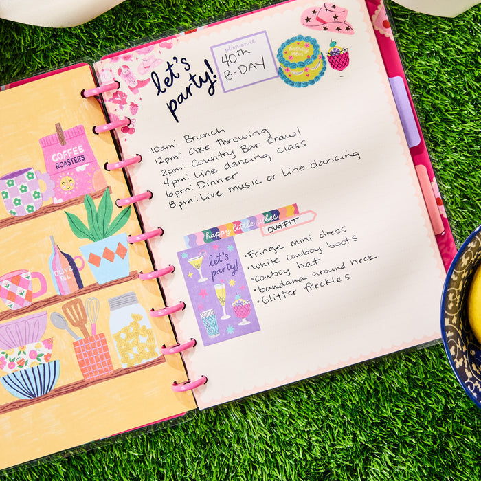 Open Happy Little Vibes - Dotted Lined Classic Notebook - 60 Sheets showing a colorful, handwritten party planning page with stickers and drawings on grass background next to a cup of tea.