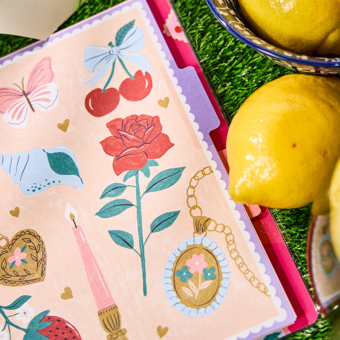 Happy Little Vibes - Dotted Lined Classic Notebook - 60 Sheets featuring a colorful cover with illustrations of a rose, cherries, butterfly, candle, heart, and locket placed next to fresh lemons on a grass surface.