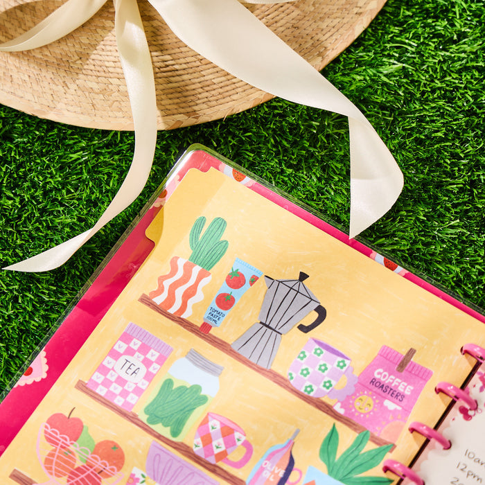 Close-up of a colorful open planner page with illustrated kitchen shelf items, next to a straw hat, featuring the Happy Little Vibes - Dotted Lined Classic Notebook - 60 Sheets.