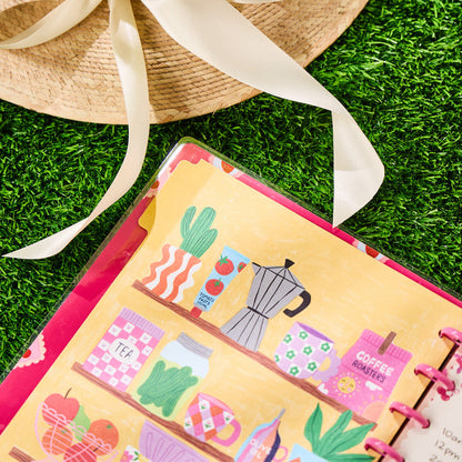 Close-up of a colorful open planner page with illustrated kitchen shelf items, next to a straw hat, featuring the Happy Little Vibes - Dotted Lined Classic Notebook - 60 Sheets.