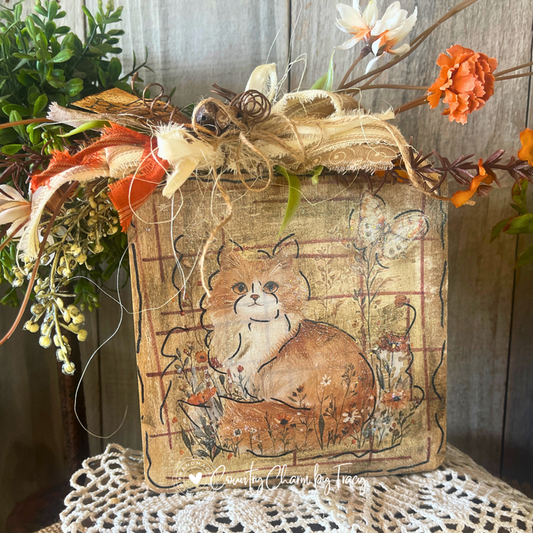 Close up of Hey Pumpkin Fluffy Orange Cat Wood Block Decor showing hand-lettered hey pumpkin distressed tag rusty jingle bell cluster layered cream lace and orange ribbon bow with wispy raffia by Country Charm by Tracy