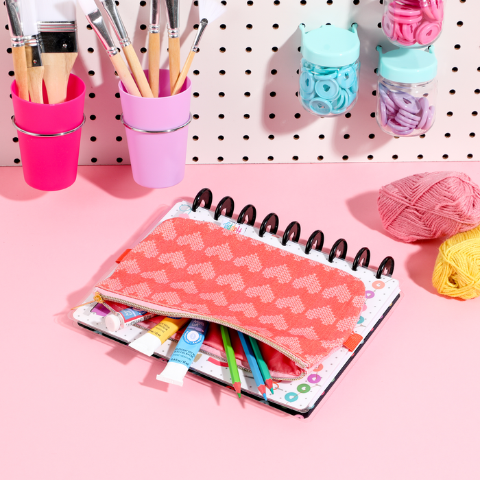 Miss Maker - Classic Banded Pen Pouch in coral with heart pattern holding colored pencils and paint tubes on a disc bound planner, surrounded by art supplies and yarn on a pink surface.
