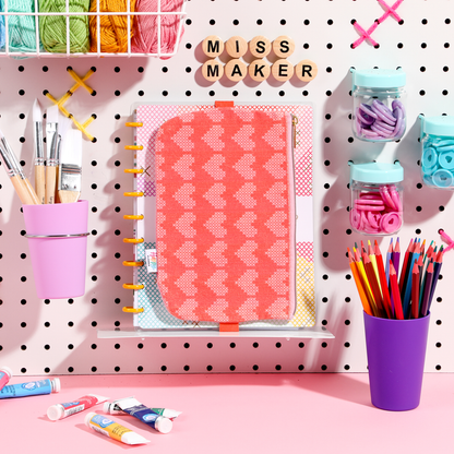 Miss Maker - Classic Banded Pen Pouch in coral pink with a heart pattern, displayed on a pegboard with colored pencils, paintbrushes, and craft supplies for planners and journals.