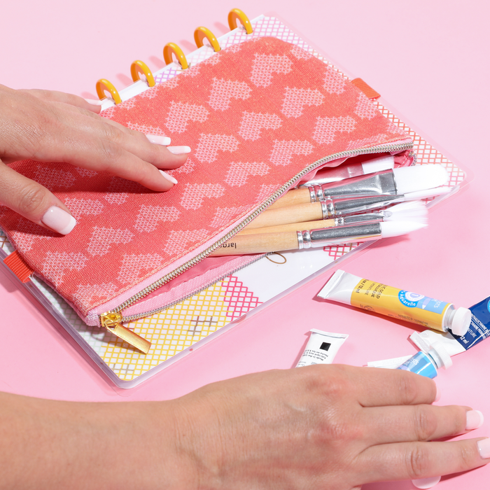 Hand holding a pink and orange Miss Maker - Classic Banded Pen Pouch unzipped, revealing paintbrushes inside, placed on a disc-bound planner with paint tubes nearby on a pink surface.