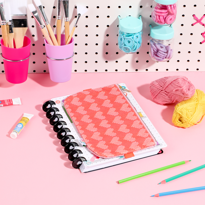 Miss Maker - Classic Banded Pen Pouch in coral with heart pattern rests on a disc-bound planner surrounded by colored pencils, paintbrushes, and yarn on a pink workspace.