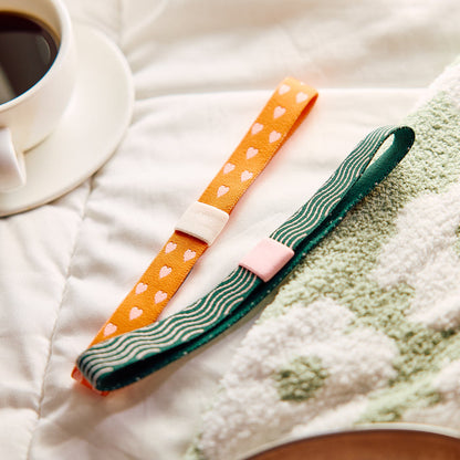Screen Free Society - Elastic Band Pen Holder - 2 Pack with orange heart and green wave designs displayed on a cozy bed beside coffee