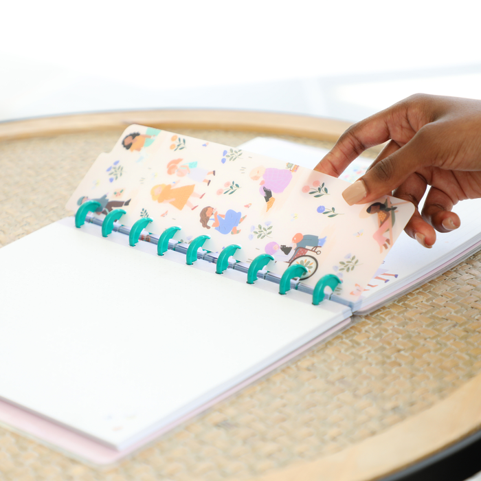 Hand holding a decorative translucent page with illustrated people flipping through a disc-bound planner on a woven surface, featuring the Squad Goals Love Every Season - Classic Accessory Pack.