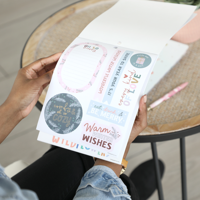 Person holding a sheet of stickers featuring seasonal and cozy phrases like "eat, drink & be merry" and "Warm Winter Wishes" from the Squad Goals Love Every Season - Large Value Pack Stickers collection, with a planner and pen on a table nearby.