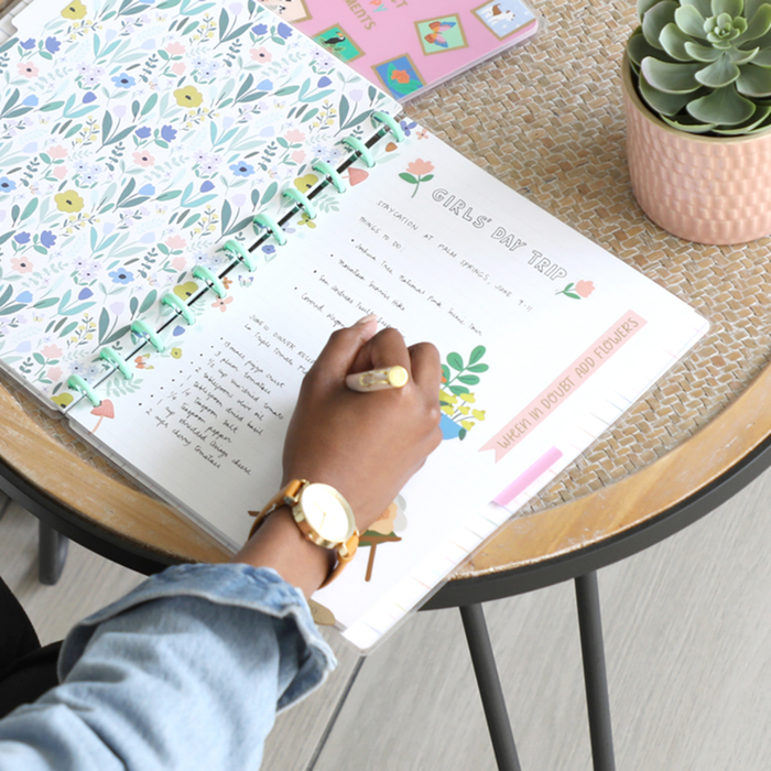 Person writing in a disc bound planner decorated with floral designs and stickers, featuring "Squad Goals Love Every Season - Large Value Pack Stickers" on a wooden table next to a succulent plant.