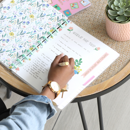 Person writing in a disc bound planner decorated with floral designs and stickers, featuring "Squad Goals Love Every Season - Large Value Pack Stickers" on a wooden table next to a succulent plant.